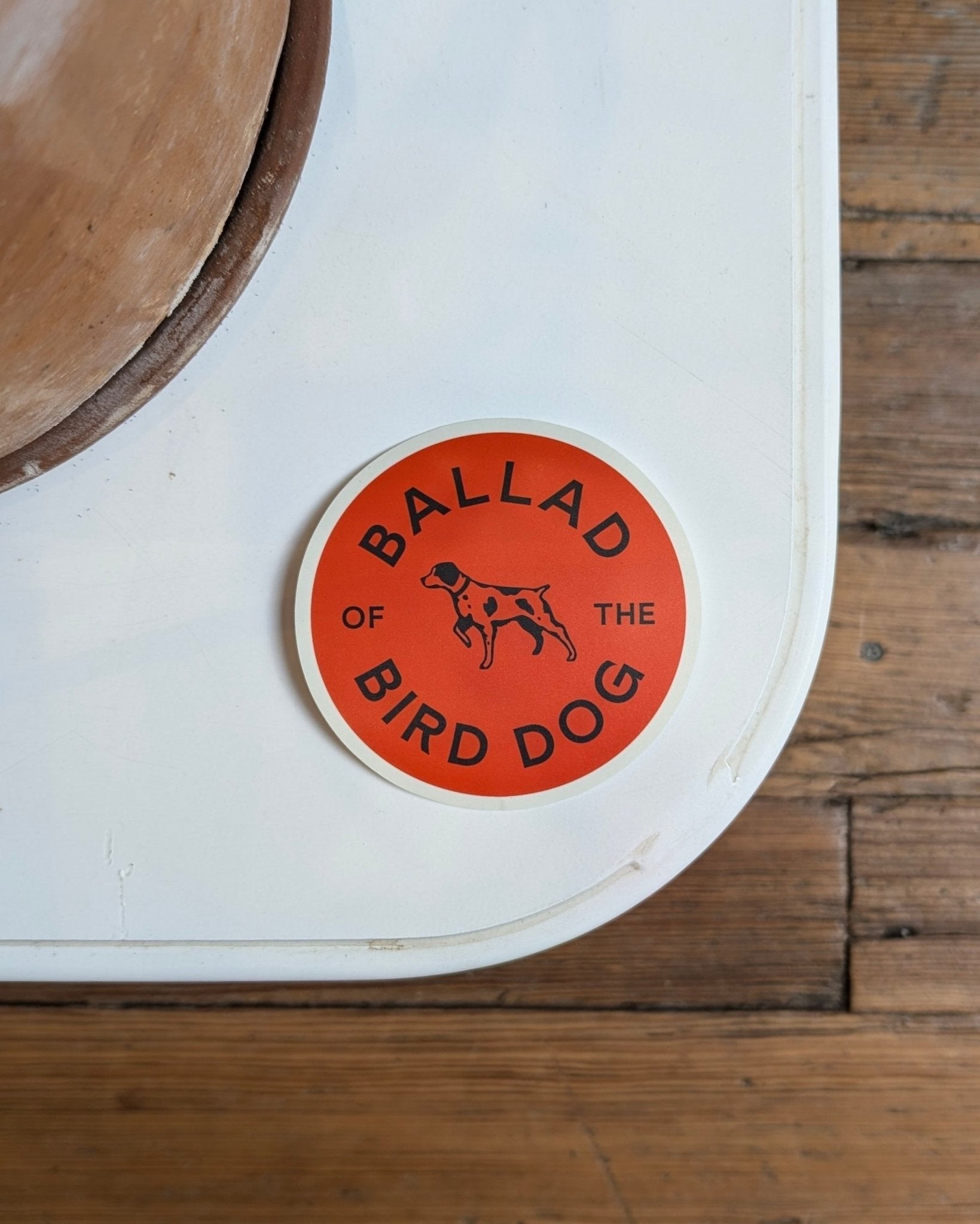 The Ballad of the Bird Dog Shop Sticker features an orange bird dog design—durable and perfect for coolers or outdoor gear. Shown on a white surface with wood flooring in the background.