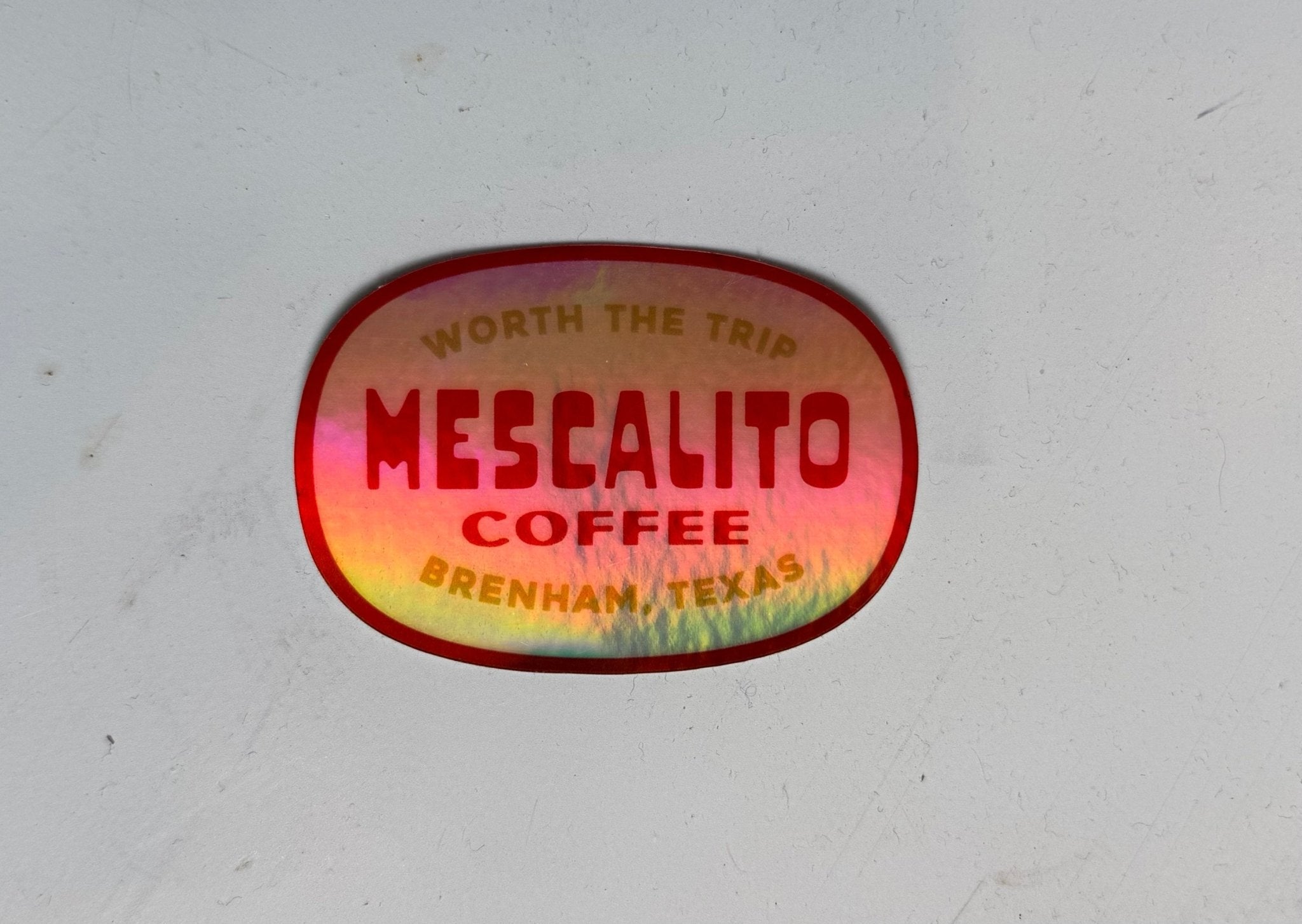 A glossy red Shop Sticker from Mescalito Coffee reads "Mescalito Coffee, Brenham, Texas. Worth the trip" on a light gray surface—celebrating this inviting small-town coffee shop.