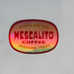 A glossy red Shop Sticker from Mescalito Coffee reads "Mescalito Coffee, Brenham, Texas. Worth the trip" on a light gray surface—celebrating this inviting small-town coffee shop.