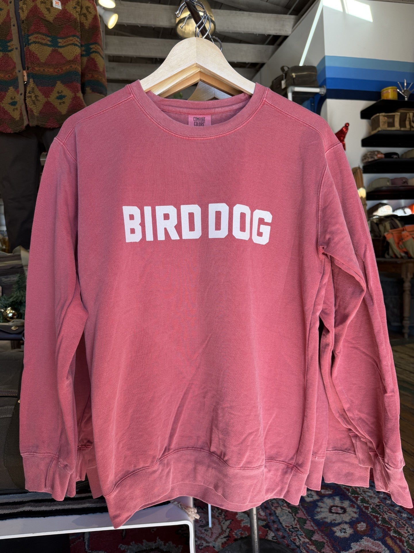 The Ballad of the Bird Dog Shop Sweater, also called the Bird Dog Honor Crewneck, is a red vintage-washed crewneck with bold white "BIRD DOG" lettering, shown displayed on a hanger in a clothing store.
