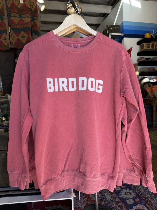The Ballad of the Bird Dog Shop Sweater, also called the Bird Dog Honor Crewneck, is a red vintage-washed crewneck with bold white "BIRD DOG" lettering, shown displayed on a hanger in a clothing store.