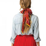 A woman with long, wavy hair is seen from the back wearing a Jenni Earle Silk Twilly Scarf “Giddy Up,” featuring horseshoe patterns tied in a bow, paired with a light denim shirt and red pleated skirt against a white background.