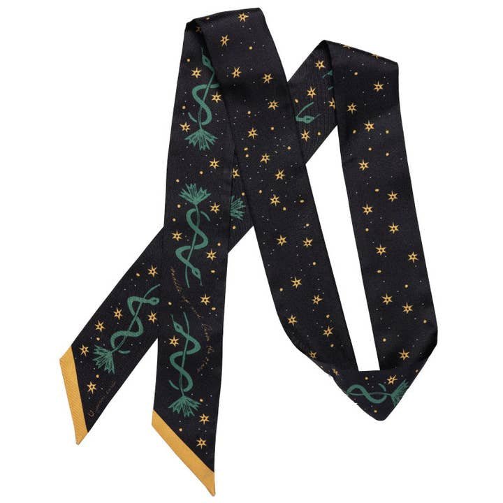 The Jenni Earle “Saddle Up and Lasso the Star” Silk Twilly Scarf features gold stars, green caduceus designs, and yellow-tipped ends—adding a touch of celestial luxury to any outfit.
