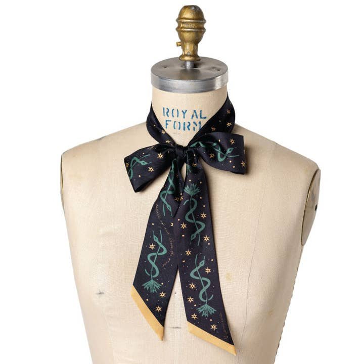 The Jenni Earle “Saddle Up and Lasso the Star” Silk Twilly Scarf features green snake and star motifs, elegantly bow-tied around a dress form and crafted from lustrous black silk.