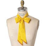 A dress form showcases the Jenni Earle Silk Twilly Scarf, “This Ain't My First Rodeo,” tied in a bow at the neck, highlighting its yellow hue, subtle pattern, and decorative edges.
