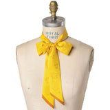 A dress form showcases the Jenni Earle Silk Twilly Scarf, “This Ain't My First Rodeo,” tied in a bow at the neck, highlighting its yellow hue, subtle pattern, and decorative edges.