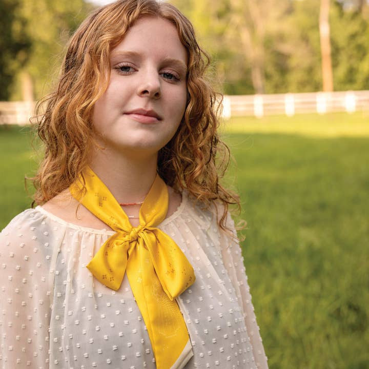 A young person with long curly hair, wearing a white textured blouse and the Jenni Earle “This Ain't My First Rodeo" Silk Twilly Scarf, stands outdoors in a grassy area with a white fence in the background.