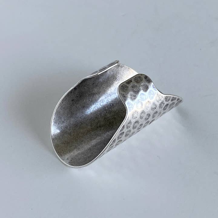 The Silver Cigar Band Ring by Ornamental Things, featuring a curved and textured hammered silver finish, rests on a white surface.