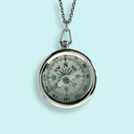 The Silver Compass Necklace by Ornamental Things features a working silver compass pendant on a long chain, shown against a light blue background.
