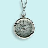 The Silver Compass Necklace by Ornamental Things features a working silver compass pendant on a long chain, shown against a light blue background.