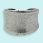 The Silver Concave Ring by Ornamental Things features a simple, unadorned wide band of silver-toned metal with a gentle curve, displayed on a light blue background.