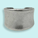 The Silver Concave Ring by Ornamental Things features a simple, unadorned wide band of silver-toned metal with a gentle curve, displayed on a light blue background.