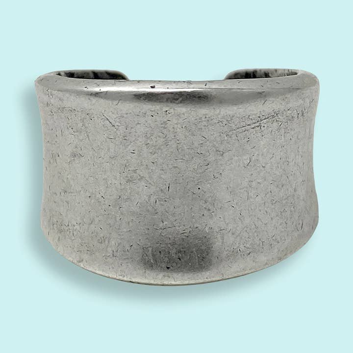 The Silver Concave Ring by Ornamental Things features a simple, unadorned wide band of silver-toned metal with a gentle curve, displayed on a light blue background.