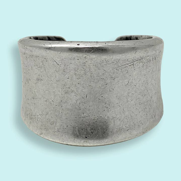 A wide, matte silver cuff bracelet is displayed on a light blue background, paired elegantly with the Silver Concave Ring by Ornamental Things.