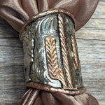 The Silver Engraved Coyote N' Cactus Wild Rag Slide by Rodeo Drive features engraved cactus designs in silver and copper tones, shown on a brown fabric napkin atop a wooden surface.