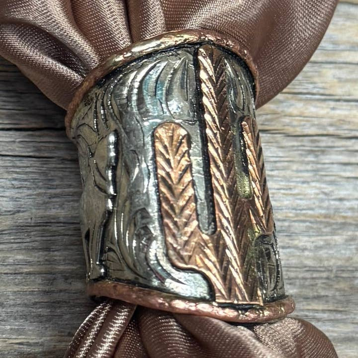 Close-up of the Rodeo Drive Silver Engraved Coyote N' Cactus Wild Rag Slide, featuring cactus and wheat designs, holding a brown satin napkin on a wooden surface.