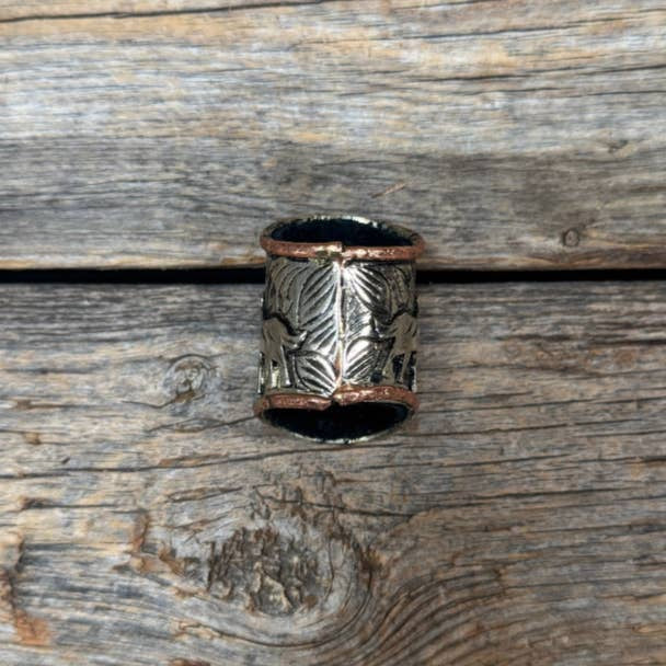 The Silver Engraved Coyote N' Cactus Wild Rag Slide by Rodeo Drive, featuring leaf engravings and copper-toned edges, is displayed on a weathered wooden surface.