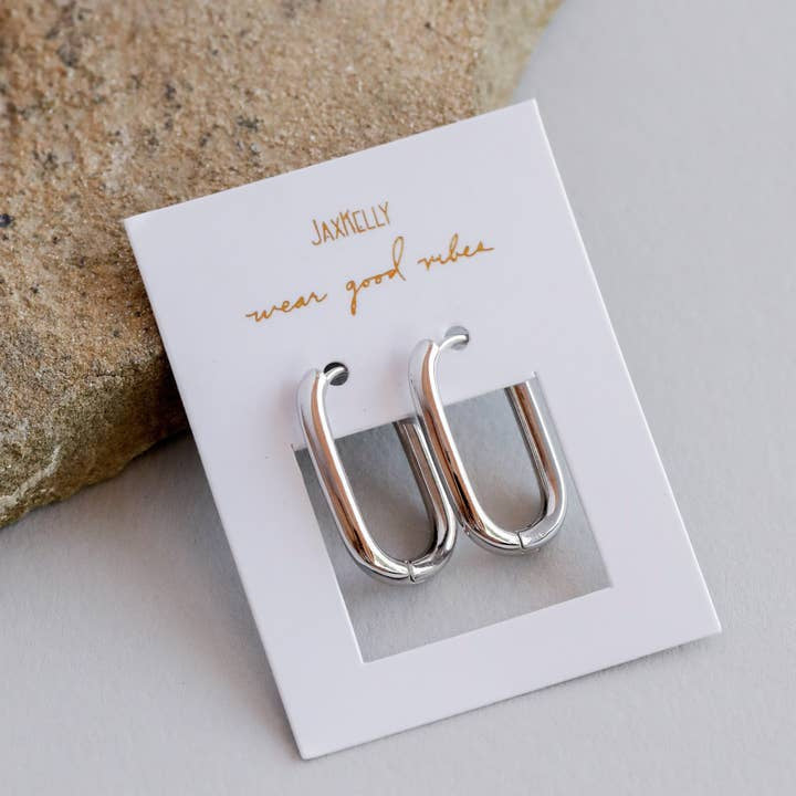 A pair of tarnish-free Silver Hoop - Large Rectangle Earrings by JaxKelly, displayed on a white "JaxKelly wear good vibes" card next to a stone surface.
