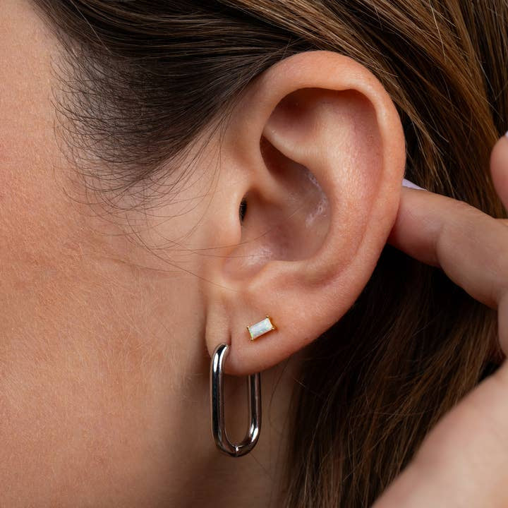 A hand gently touches an ear adorned with JaxKelly's Silver Hoop - Large Rectangle Earrings, featuring a shiny rectangular stud and a large silver hoop.