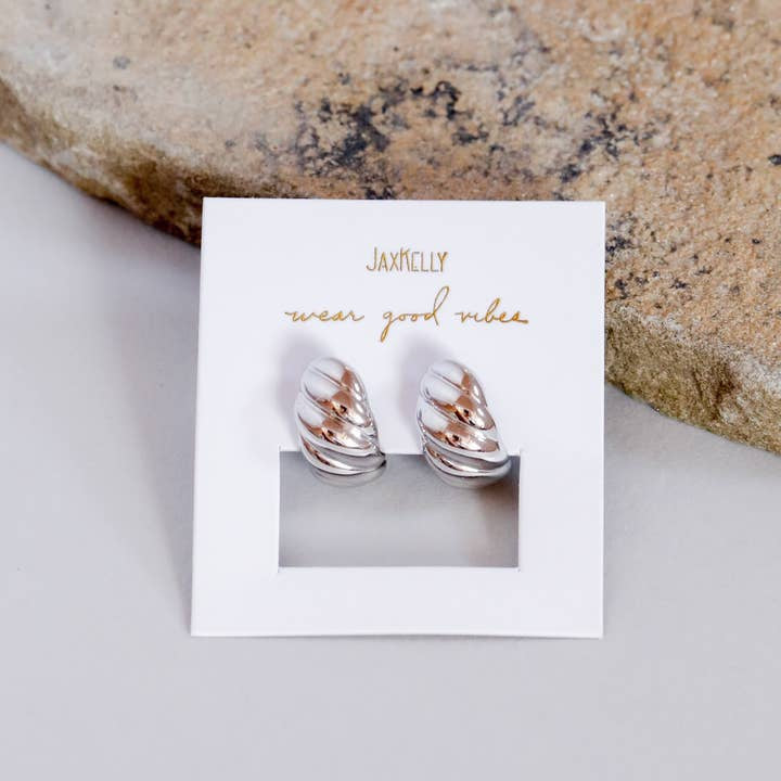 A pair of hypoallergenic, tarnish-free Silver Hoop Swirl Earrings by JaxKelly is displayed on a white "JaxKelly wear good vibes" card next to a textured stone.