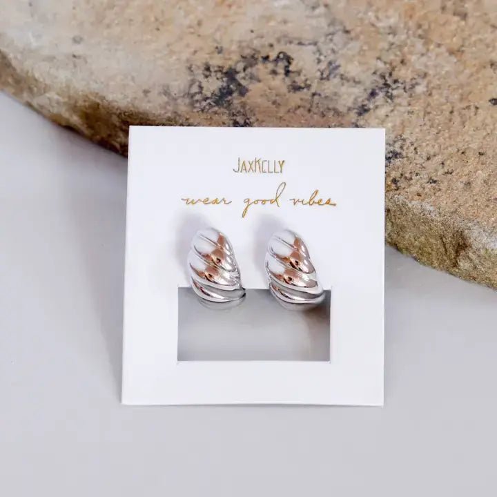 A pair of silver swirl hoop earrings from JaxKelly, displayed on a “wear good vibes” branded card, set against a mottled stone background.