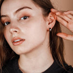 A person with long brown hair touches their ear, showcasing the JaxKelly Silver Hoop - Wide - Earrings by JaxKelly—a small, tarnish-free silver hoop. The background is a plain light brown color.