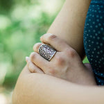 A person with arms crossed wears the Silver Leaf Lace Ring by Ornamental Things—a large, intricately patterned adjustable sterling silver ring—on their finger, set against a blurred green background.