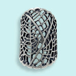 The Silver Leaf Lace Ring by Ornamental Things is a wide sterling silver ring featuring an intricate openwork lace pattern, shown on a light blue background.