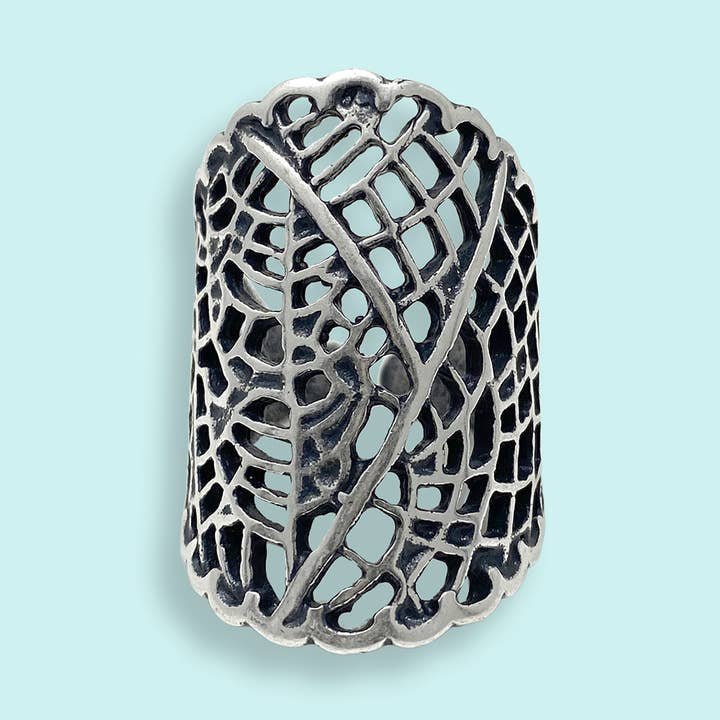 The Silver Leaf Lace Ring by Ornamental Things is a wide sterling silver ring featuring an intricate openwork lace pattern, shown on a light blue background.