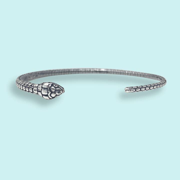 The Silver Open Snake Bangle Bracelet by Ornamental Things features detailed head and tail designs, elegantly wraps around your wrist, and is adjustable. Shown against a light blue background.