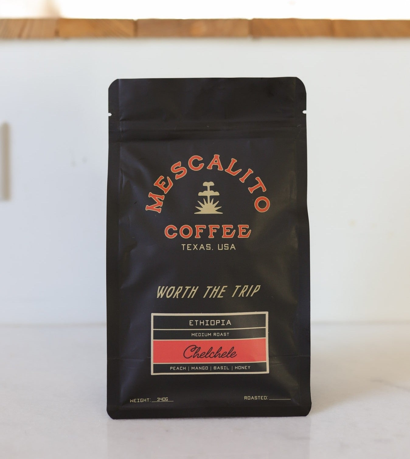 A black bag of Mescalito Coffee’s Single Origin Whole Coffee Beans | Chelchele features Ethiopia Medium Roast, crafted by the anaerobic natural process with tasting notes of peach, mango, basil, and honey and "Worth the Trip" on the label.