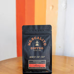 A black bag of Mescalito Coffee’s Single Origin Whole Coffee Beans from Madriz rests on a wooden table, labeled as organic with caramel, orange, melon, and apple notes—a medium roast offering a vibrant and flavorful cup.