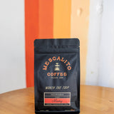 A black bag of Mescalito Coffee’s Single Origin Whole Coffee Beans from Madriz rests on a wooden table, labeled as organic with caramel, orange, melon, and apple notes—a medium roast offering a vibrant and flavorful cup.