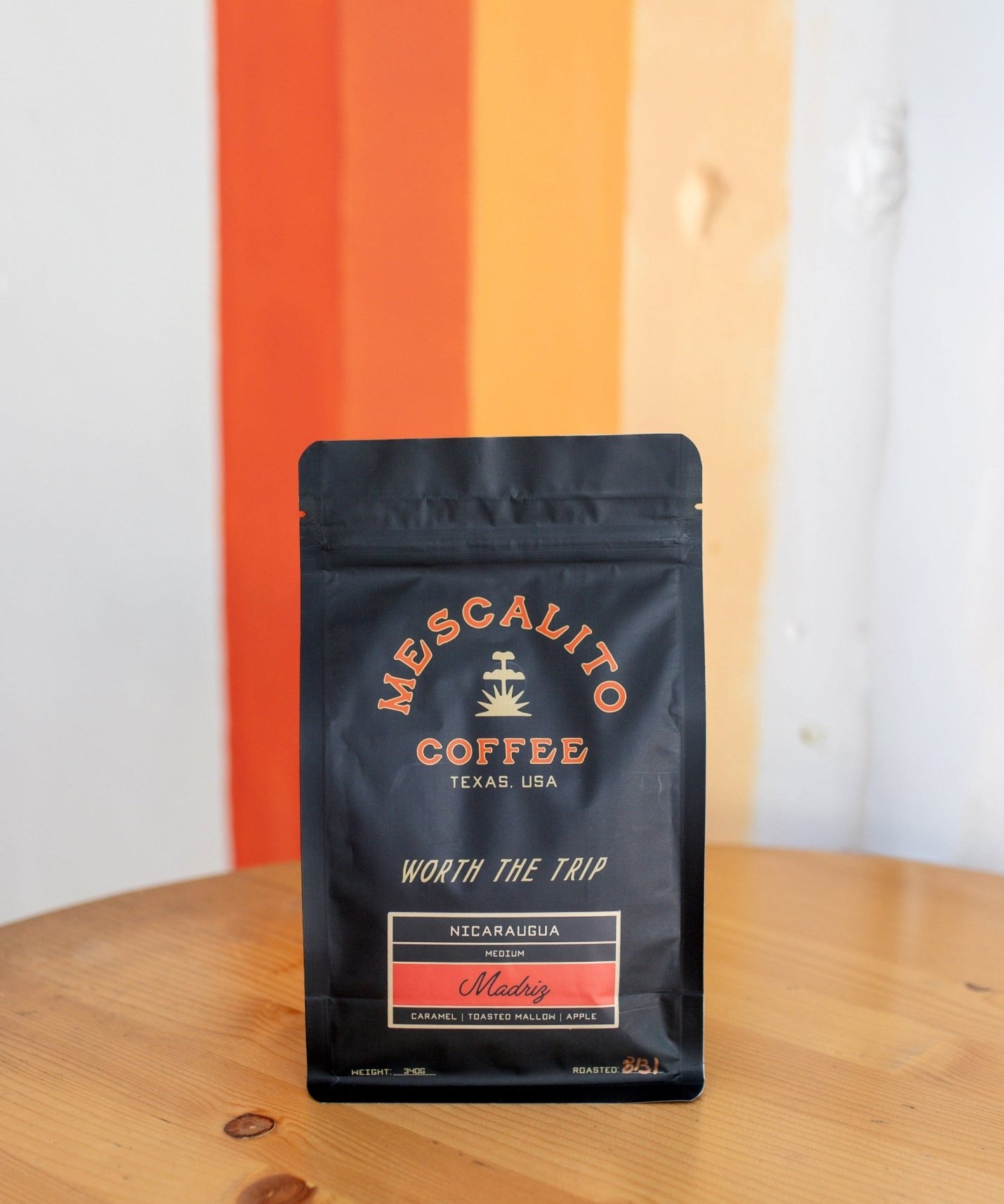 A black bag of Mescalito Coffee’s Single Origin Whole Coffee Beans from Madriz rests on a wooden table, labeled as organic with caramel, orange, melon, and apple notes—a medium roast offering a vibrant and flavorful cup.