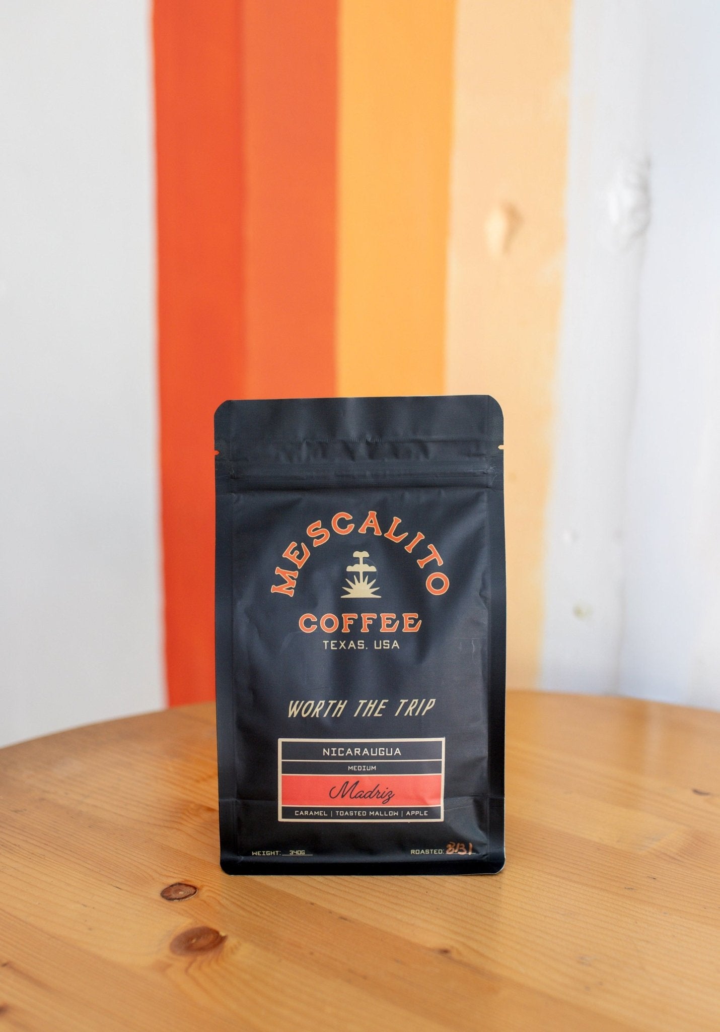 A black bag of Mescalito Coffee’s Single Origin Whole Coffee Beans from Madriz rests on a wooden table, labeled as organic with caramel, orange, melon, and apple notes—a medium roast offering a vibrant and flavorful cup.