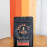 A black bag of Mescalito Coffee’s Single Origin Whole Coffee Beans | Magnolia rests on a wooden table before an orange and white striped wall, labeled as Guatemalan whole bean coffee with a washed process and medium roast.