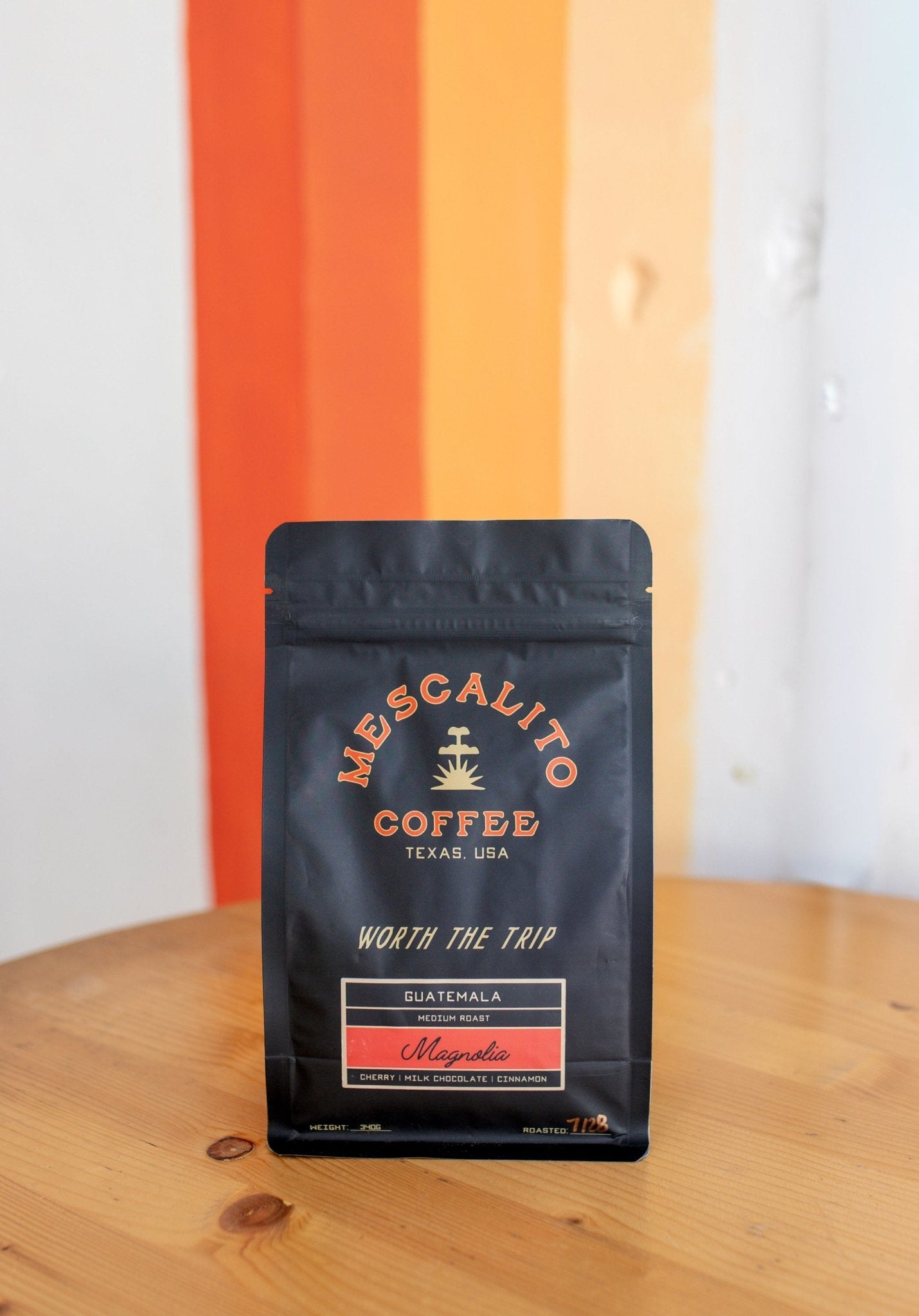 A black bag of Mescalito Coffee’s Single Origin Whole Coffee Beans | Magnolia sits on a wooden table against an orange and white striped wall, featuring this medium roast from Guatemala.