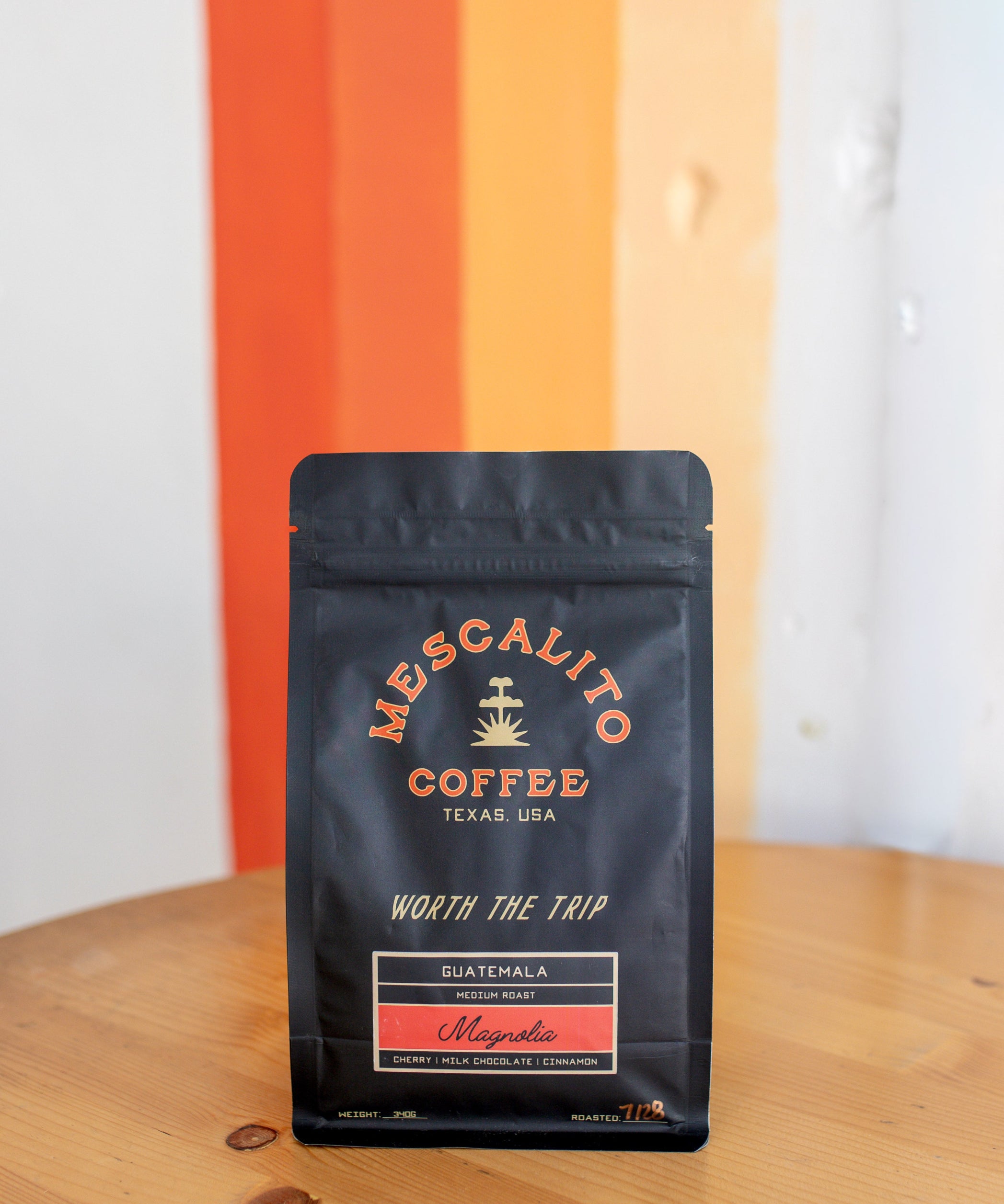 A black bag of Mescalito Coffee’s Single Origin Whole Coffee Beans | Magnolia rests on a wooden table before an orange and white striped wall, labeled as Guatemalan whole bean coffee with a washed process and medium roast.