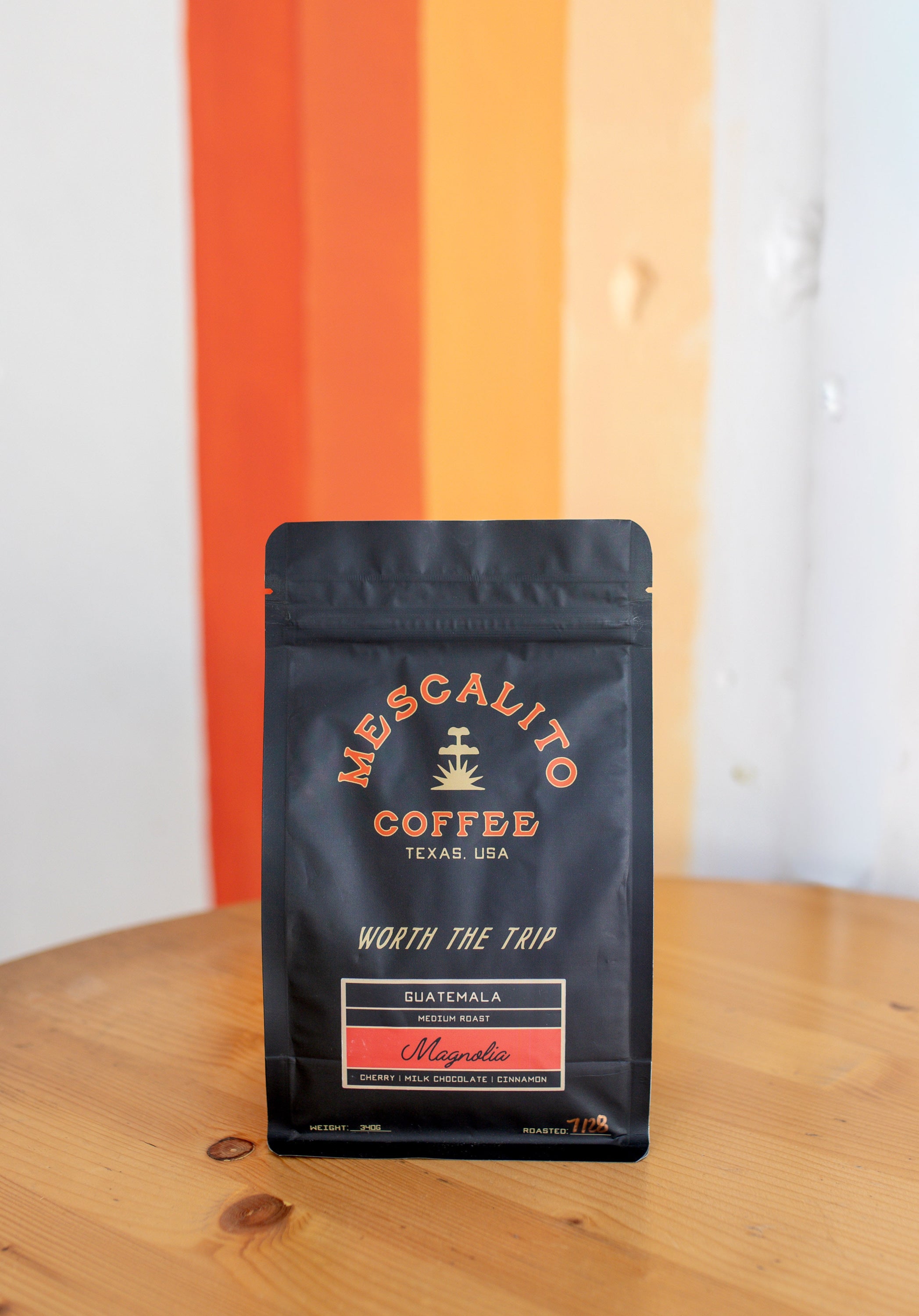 A black bag of Mescalito Coffee’s Single Origin Whole Coffee Beans | Magnolia rests on a wooden table before an orange and white striped wall, labeled as Guatemalan whole bean coffee with a washed process and medium roast.