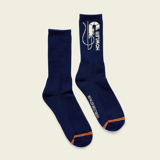 A pair of navy blue Skrimp crew socks by Howler Brothers, accented with orange and featuring a white graphic of a person fishing and "Howler Brothers" text.