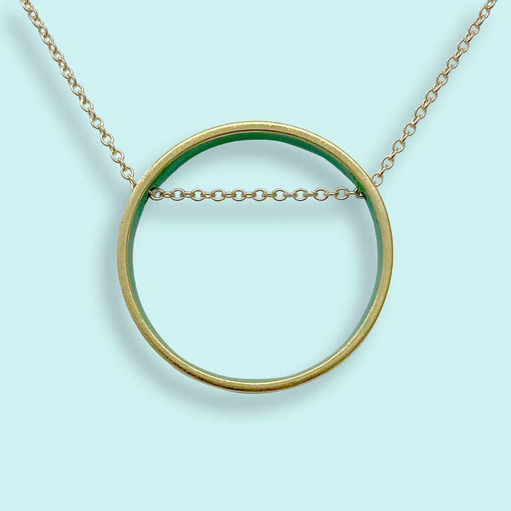 Small emerald green circle pendant necklace by Ornamental Things with a cable chain passing through the center, displayed on a light blue background.