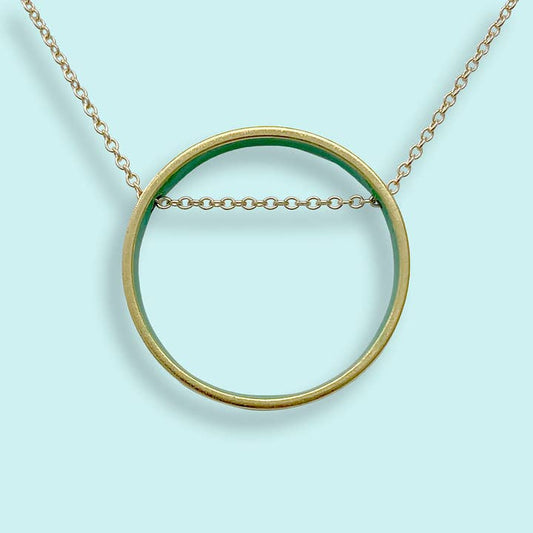 Small emerald green circle pendant necklace by Ornamental Things with a cable chain passing through the center, displayed on a light blue background.