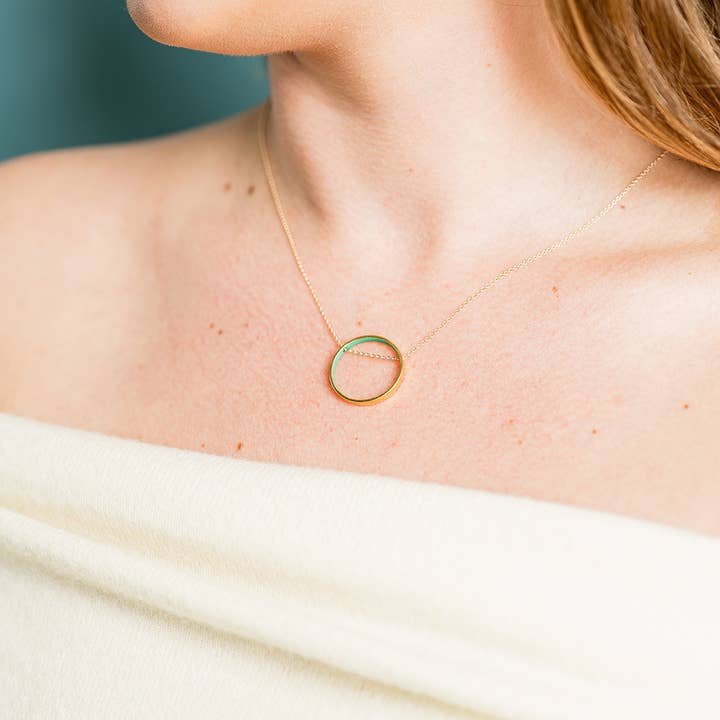 A close-up of a woman wearing the Small Emerald Green Circle Necklace by Ornamental Things, featuring a minimalist gold circle pendant with emerald enamel on a gold-filled chain, styled with an off-the-shoulder cream top.