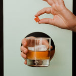 A hand holds a glass of orange liquid with ice, while another presents a Smoked Old Fashioned Cocktail Cube by Yes Cocktail Co.—perfect for crafting the ultimate Hickory smoked Old Fashioned.
