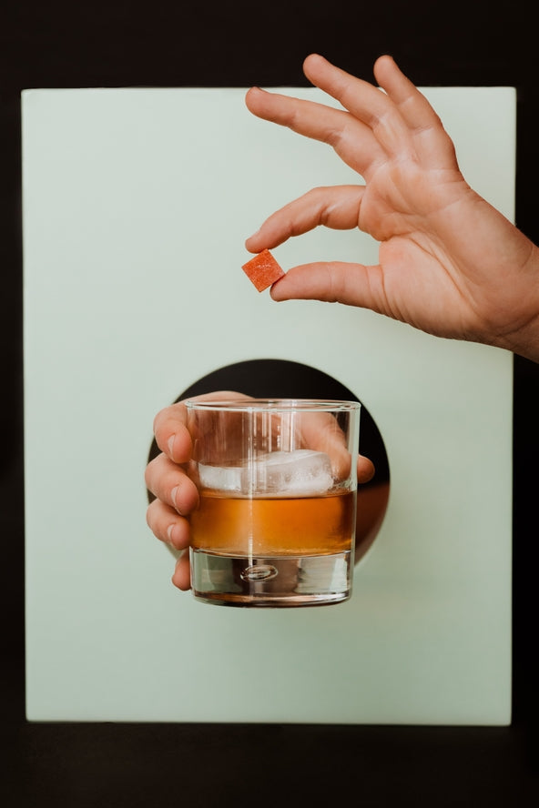 A hand holds a glass of orange liquid with ice, while another hand holds a Smoked Old Fashioned Cocktail Cube by Yes Cocktail Co. above it, set against a light green and black background.