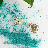 The Sommar Layered Studs | Aventurine by Peter and June, featuring gold sunburst design with green stones, rest on a white surface dusted with turquoise powder beside a green leaf.