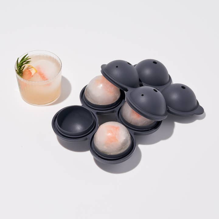A cocktail with rosemary garnish sits beside the W&P Sphere Ice Mold, a black silicone tray holding three slow-melting ice spheres—two partially covered and one uncovered.