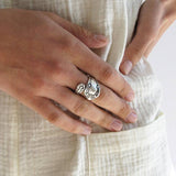 A hand rests on cream fabric, wearing the Ornamental Things Spoon Ring—an adjustable, vintage-inspired ring crafted from sterling silver plate with ornate detailing.