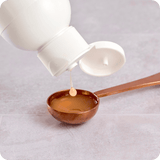 A white bottle dispenses liquid into the Stainless Steel Serving Spoon by Rho Nutrition, resting on a light surface.