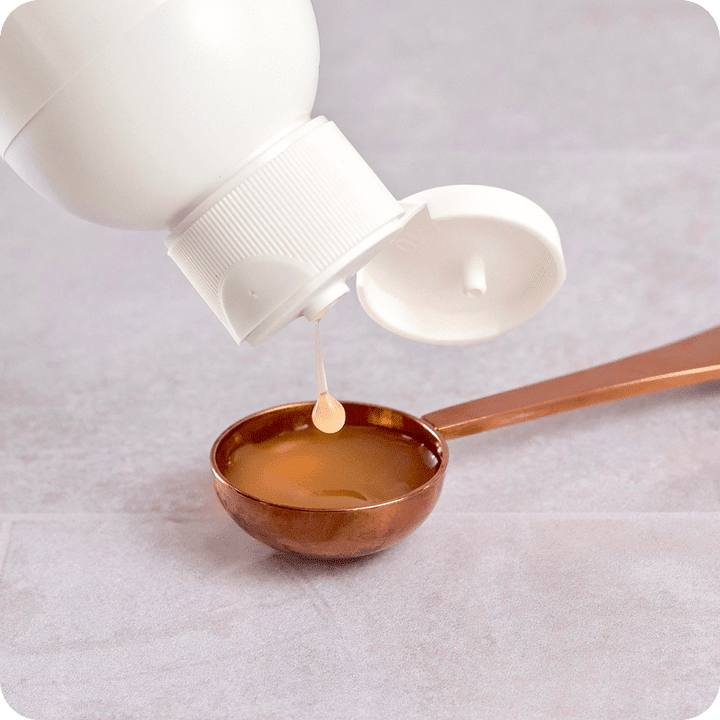 A white bottle dispenses liquid into the Stainless Steel Serving Spoon by Rho Nutrition, resting on a light surface.